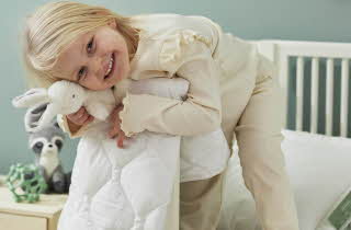 En liten jente som står på en seng og holder en kaninbamse og en dyne