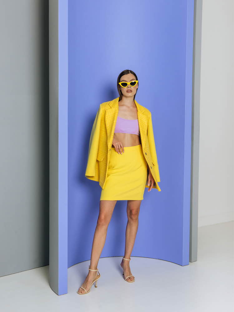 Modell med gul jakke og gult skjørt og lilla topp foran lilla vegg.