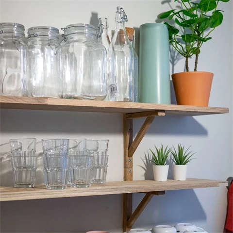 Trehvit vegghengt hylle på grønn vegg med diverse glass og kopper.