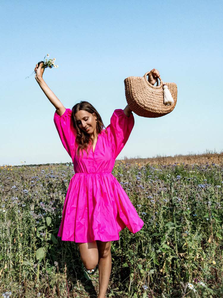Jente i knallrosa kjole poserer med veske og blomster i blomstereng