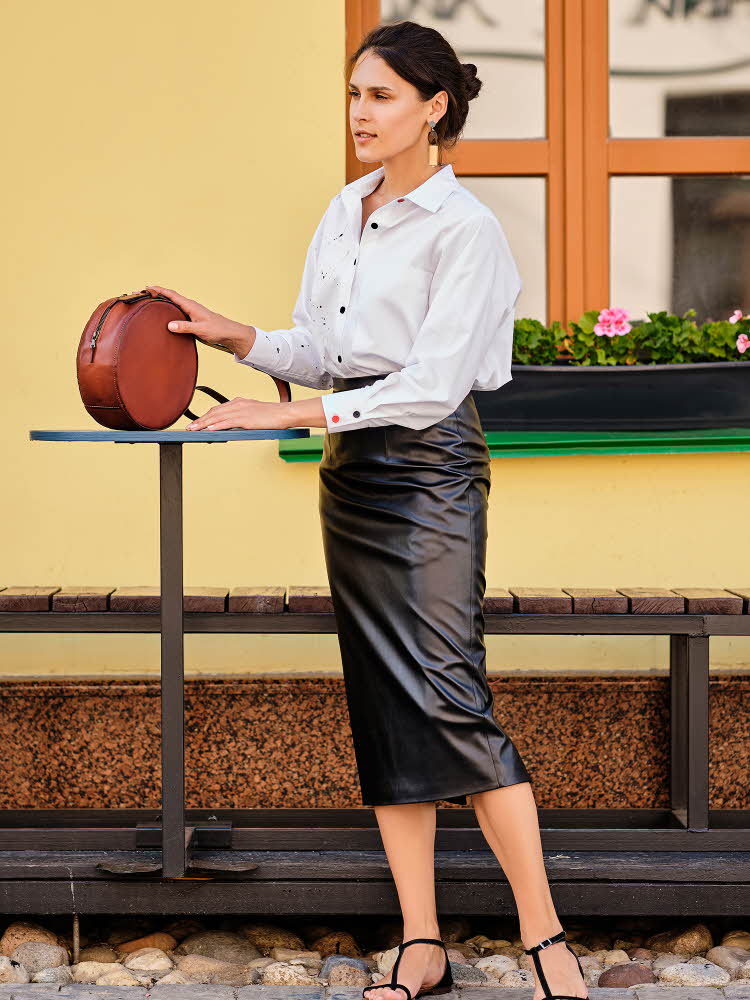 Dame med sort skinnskjørt, hvit skjorte, sorte sandaler og brun veske står ved et bord utendørs