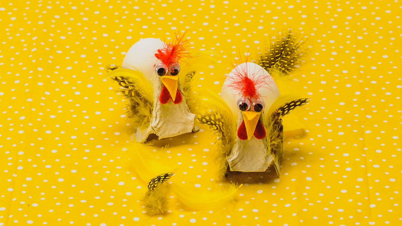 To høner, fylt med hvert sitt egg, laget av eggekartong, filtbiter og fjær på en gulprikkede bakgrunn