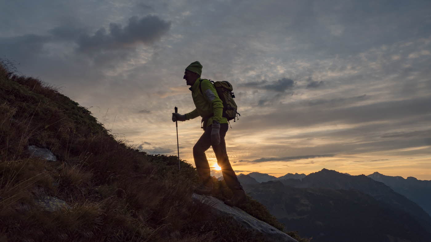 Mann går med stav i solnedgangen opp et fjell