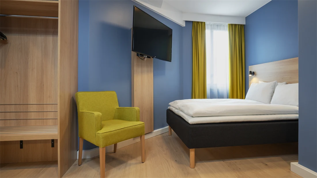 Garderobe, gul stol, blå vegger, TV og queen seng på standard rom queen på Thon Hotel Astoria.