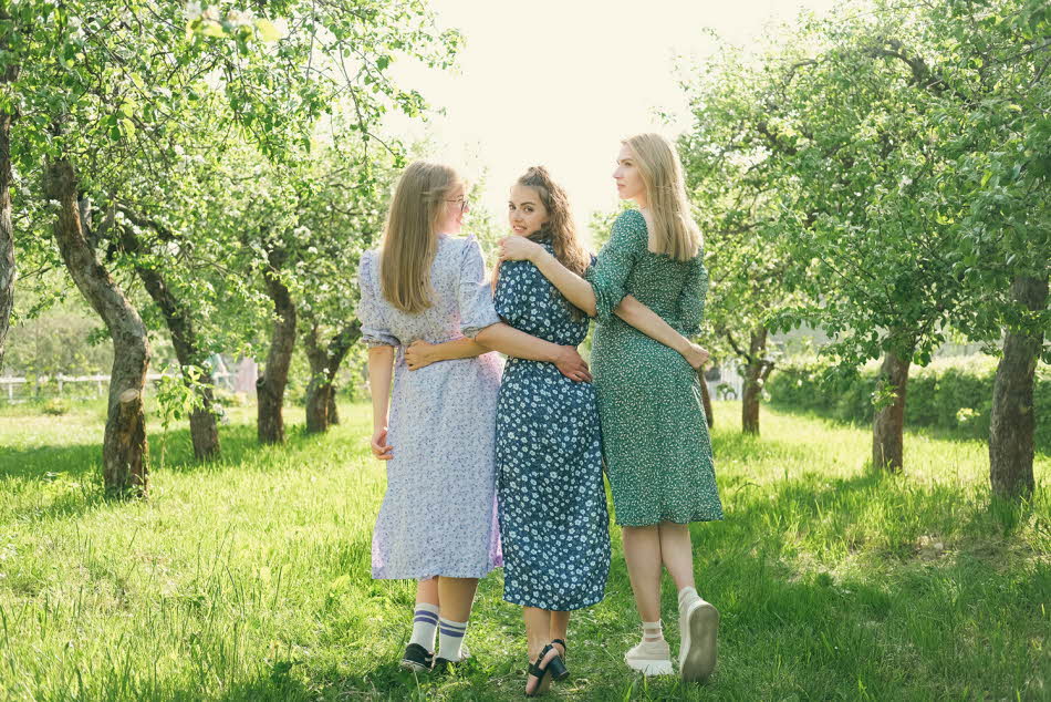 Tre jenter med blomsterkjoler holder rundt hverandre i en hage sett bakfra