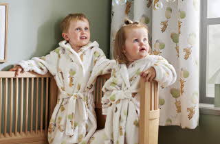 En liten gutt og jente står i like badekåper oppe i en seng