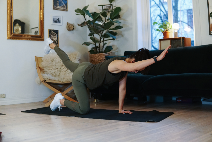 treningsekspert Anna Johansen trener på matte på gulvet, mørkegrønn sofa i bakgrunnen, lys stol med pels oppi, str grønn plante, bilder på veggen