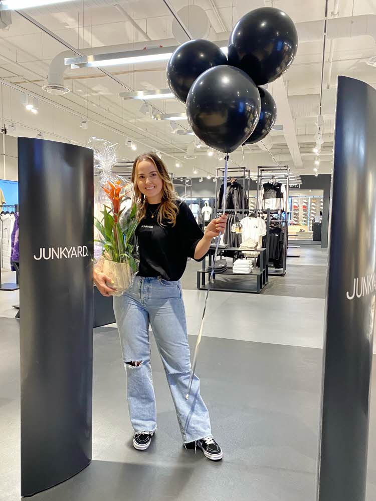 Butikksjef Helen på Junkyard holder en blomst i cellofan og en klase ballonger i butikkfasaden