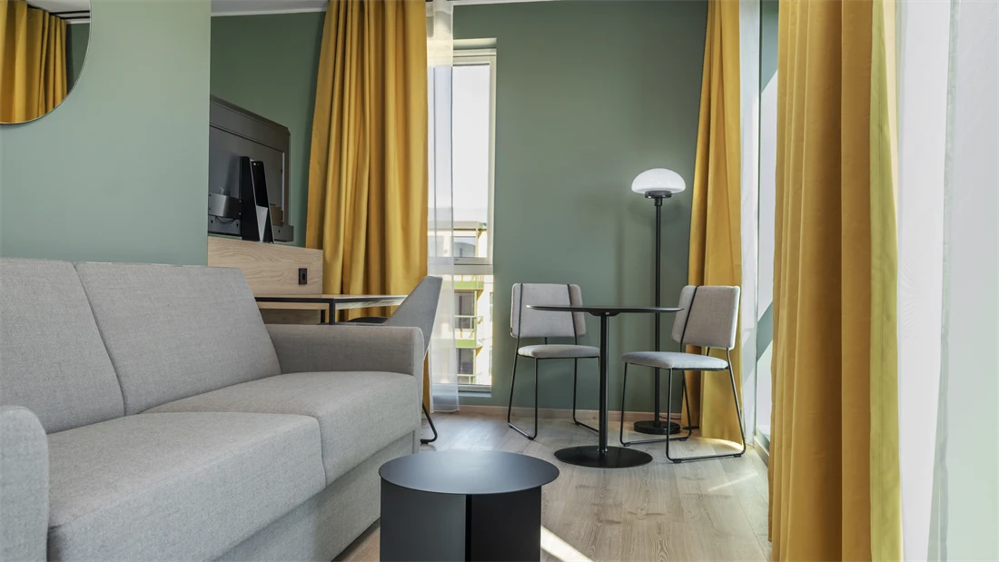Standard queen rom med grå sofa, spiseplass, vinduer, gule gardiner og grønne vegger.