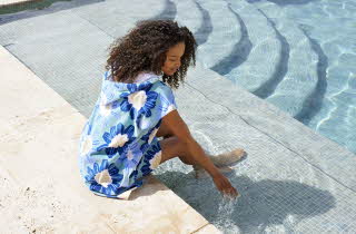 En kvinne som sitter på kanten til et basseng, hun har på seg en blå strandponcho med blå blomster på