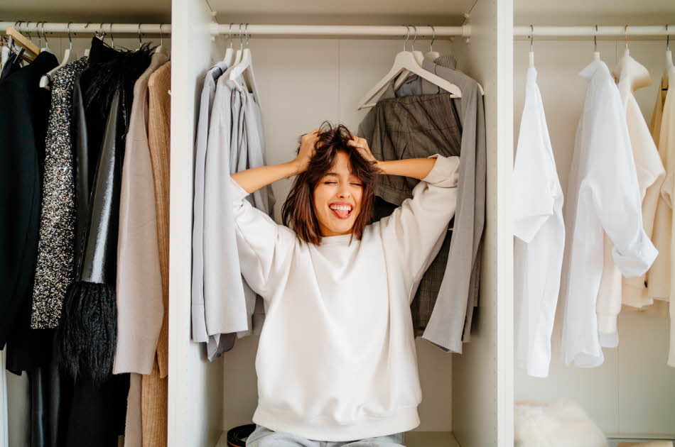 En kvinne fra livet og opp, iført hvit genser, lener seg inn i en walk-in garderobe, mens hun holder hendene i det brune håret, lukker øynene og geiper mens hun smiler.