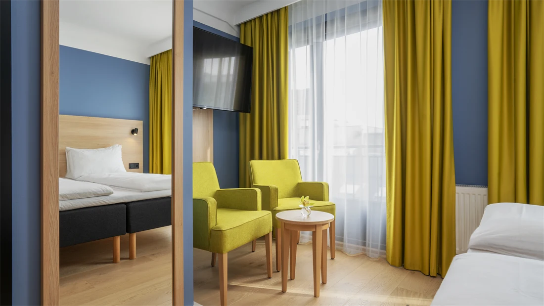 Speil, sittegruppe med to gule stoler, bord og TV, blå vegger og gule gardiner på dobbeltrom på Thon Hotel Astoria. 