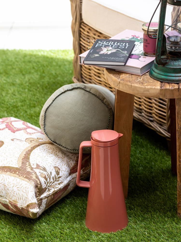 Detaljbilde, deler av kurvstol og lite trebord med bøker og lykt på kunstig gress med puter og kaffekanne