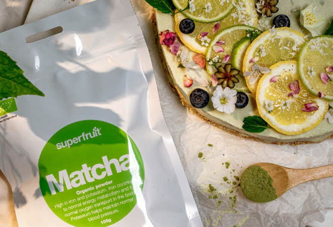 En pose med Superfruit Matcha ligger på et bord med sitron og lime og en skje med matcha pulver