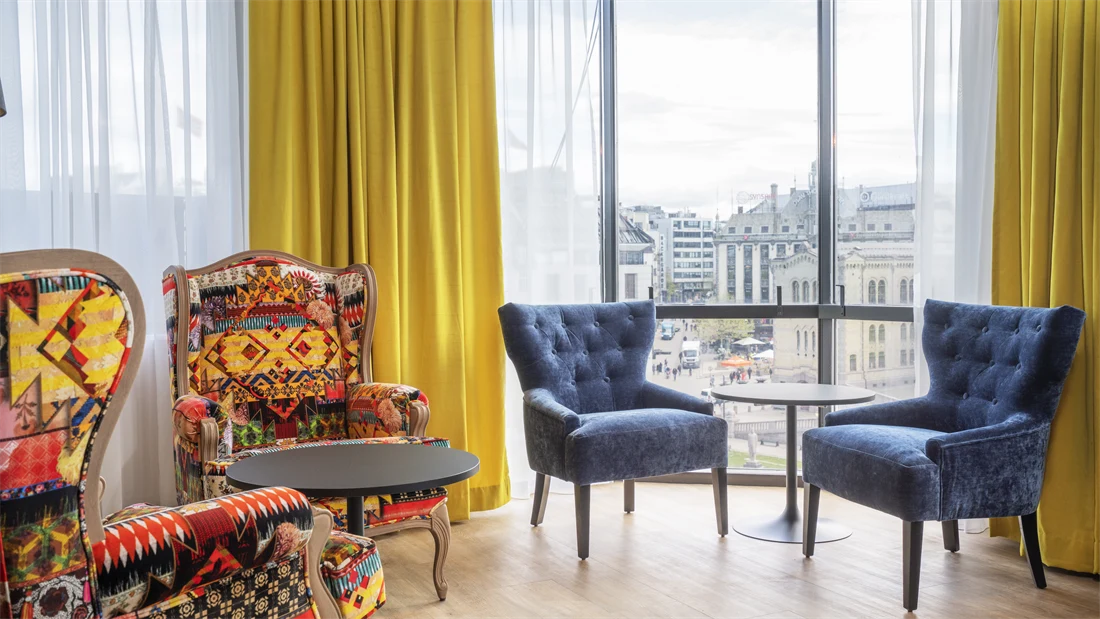 Stoler med fargerikt mønster, blå stoler, sorte runde bord, gule gardiner og store vinduer med utsikt i lounge på Thon Hotel Cecil
