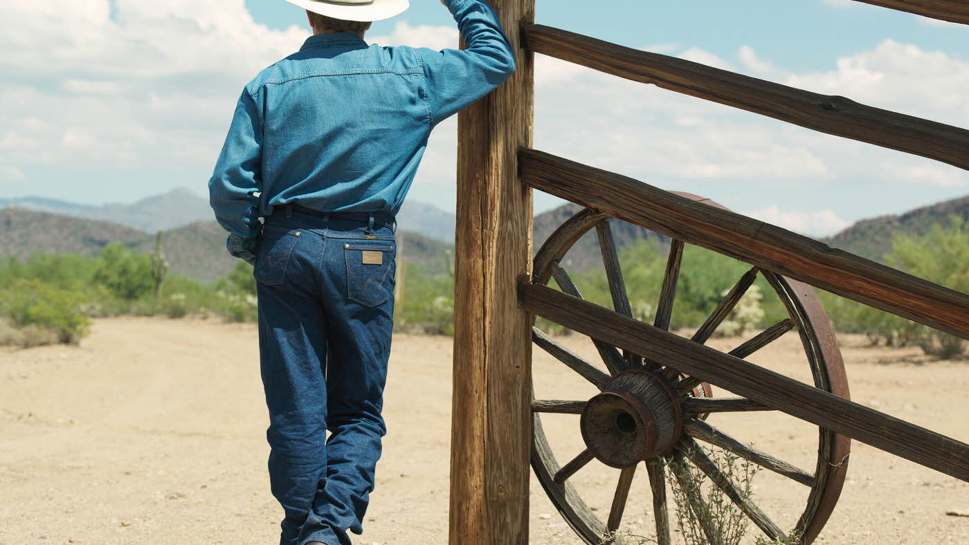 Mann i dobbel denim jeans og denimskjorte og cowboyhatt bakgra på en ranch som titter ut over landskapet.