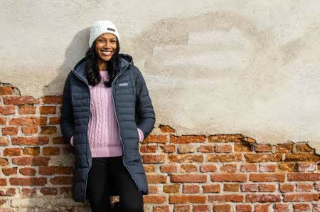 dame som smiler med hvit lue, rosa genser, lang blå høstjakke stående mot en murvegg