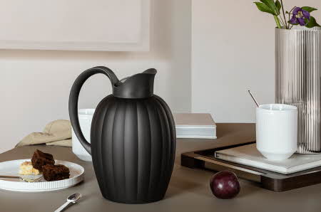 En Georg Jensen Bernadotte termokanne som står på et bord med asjett, en kopp og en vase med blomster