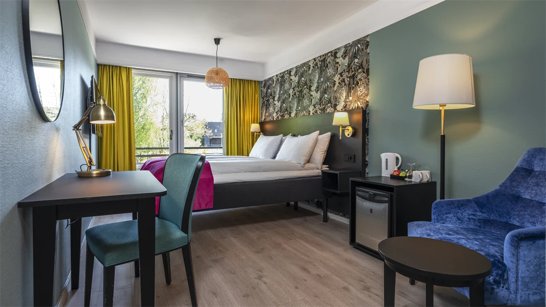 Standard rom med twin senger, rosa sengeteppe, skrivebord med tilhørende stol, gule gardiner, stort vindu og minibar.
