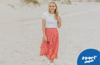 En kvinne i hvit t-skjorte og oransje blomstrete skjørt som går på en strand