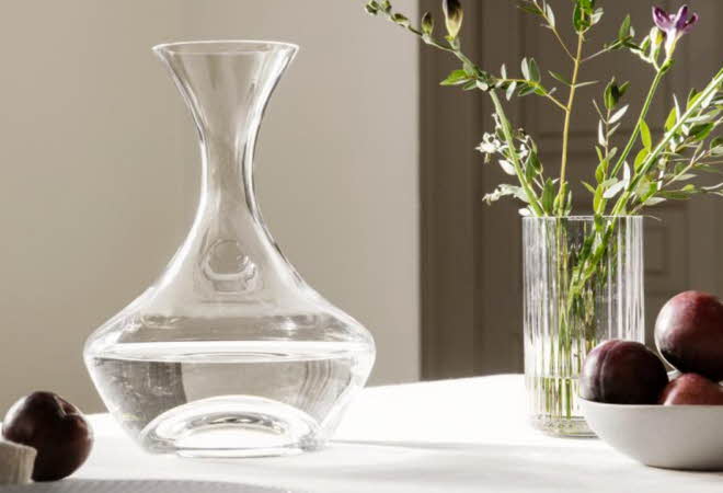 En karaffel står på et bord med en vase med blomster og en bolle med frukt.