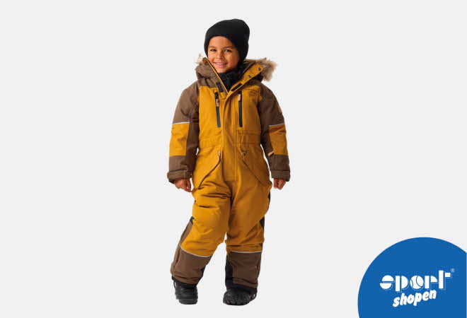 Gutt står i gul og brun vinterdress med pelskant på hetten og svarte støvler.