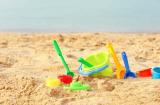Fargerike plastleker for sand, inkludert bøtter, spader og former, ligger spredt på en strand med gyllen sand og havet i bakgrunnen.