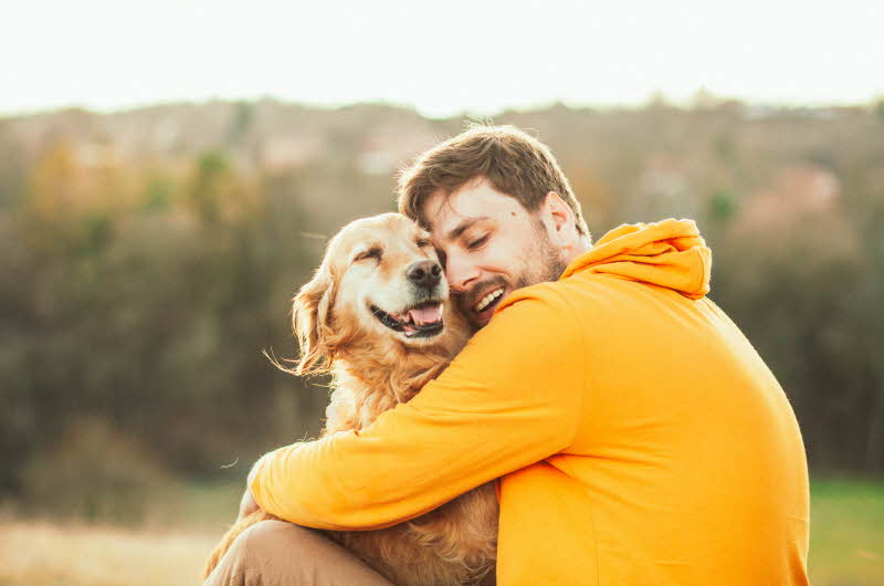Smilende kar i gul hettegenser og som med lukkede øyne koser med en smilende golden retriver hund ute i naturen