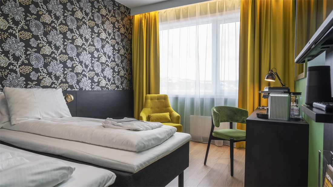 Separate senger, mørk mønstrete tapet med blomster, gule gardiner, gul stol, skrivebord med kaffemaskin og grønn stol i superior twin rom på Thon Hotel Kirkenes