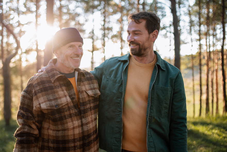 To smilende menn med rutete og grønn jakke går i skogen med trær og lysning i bakgrunnen 