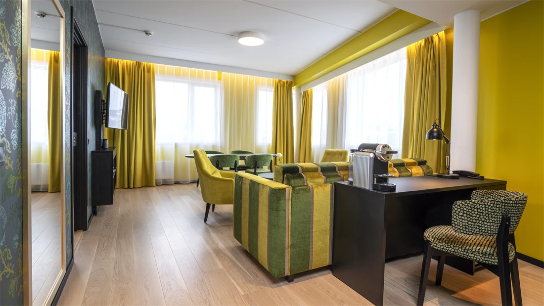 Stort speil, skrivebord med mønstrete stol, gule vegger og gul og grønn stripete sofa i suite på Thon Hotel Kirkenes