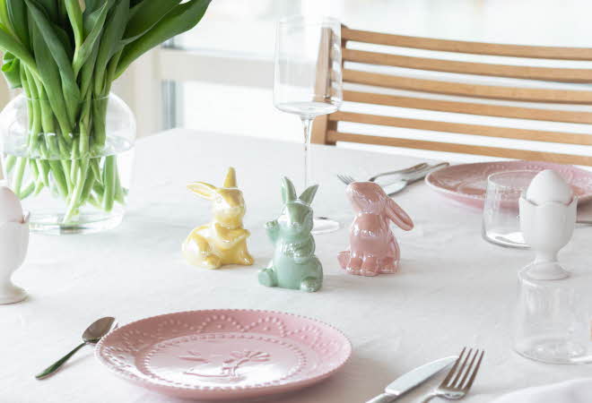 Tre kaniner i porselen står på et bord som er dekket med asjetter og bestikk.