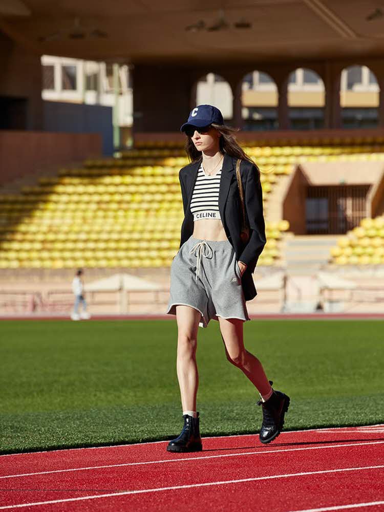Dame som går ute på en idrettsbane med grå shorts, sort dressjakke, stripete kort magetopp og blå caps