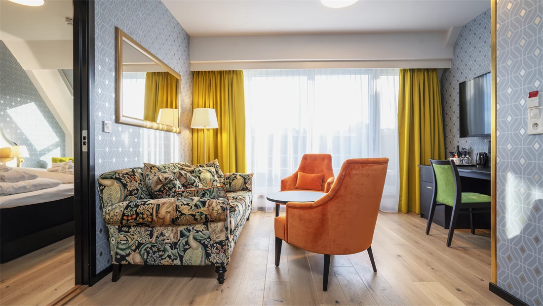 Sittegruppe med mønstret fargerik sofa, to oransje stoler, sort rundt bord og store vinduer i stue med dør åpen til soverom i suite på Thon Hotel Cecil.