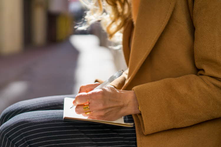 Jente som sitter ute i kamelfarget jakke og stripete bukser og skriver