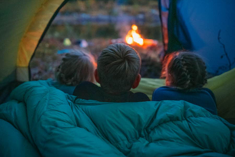 Tre barn ligger i et telt og ser ut på et bål som brenner