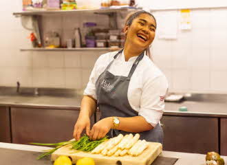 Kokken Mymint på Thon Hotel Lofoten står i et industrielt kjøkken og skjærer opp asparges, smiler og ser opp. Benk, vegg og hylle i bakgrunnen er utvidet med kunstig intelligens for å gjøre bildeformatet større enn opprinnelig.
