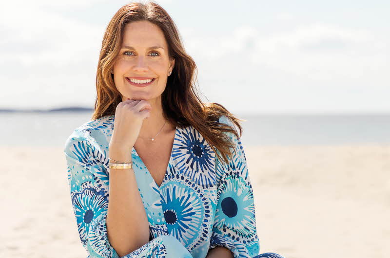 En kvinne sitter på en strand i en blå kjole med mønster, hun holder hånden under haken og smiler til kameraet