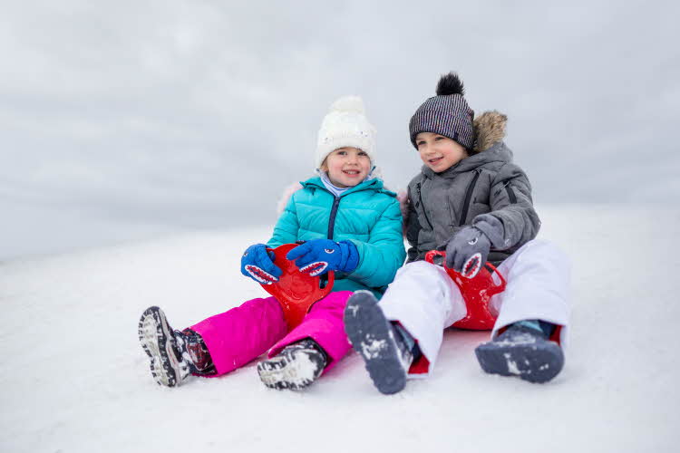 To barn sitter med akebrett på toppen av en bakke i snøen iført vintersko, boblejakker og luer.