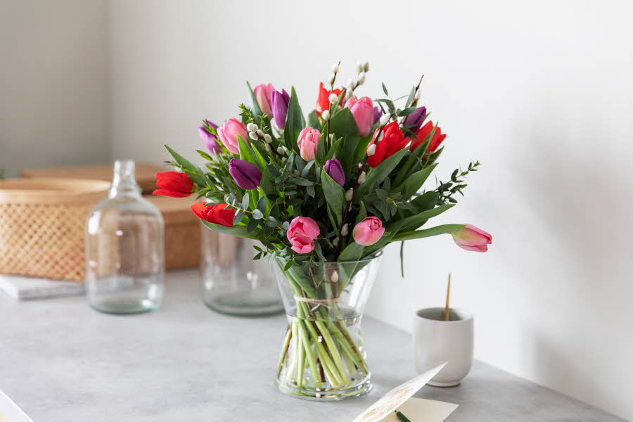 Stor bukett tulipaner står i vase på et bord, med glass og kurver bak. Noen gåsunger blant alle tulipanene i ulike farger. 