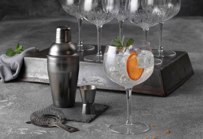 Et cocktailglass med is, appelsinskive og mynte står i sentrum, omgitt av shaker, målebeger og tomme krystallglass.