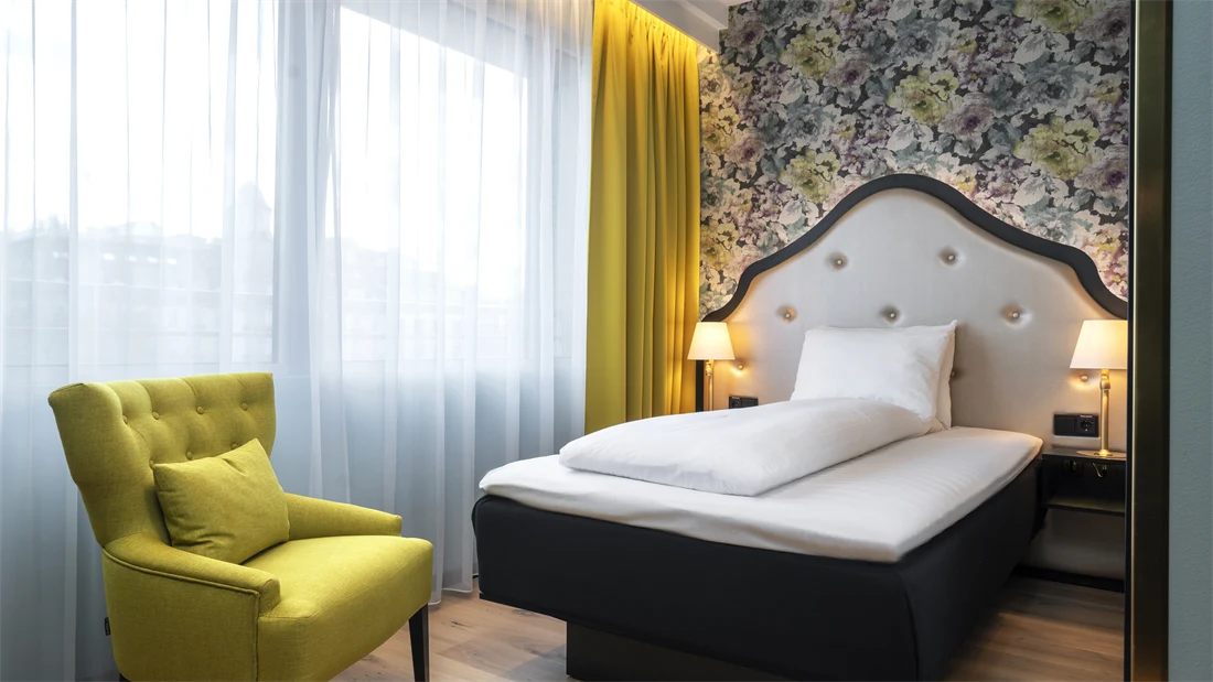 Gul stol, store vinduer, gule gardiner, blomstrete tapet og queen seng i standard rom queen på Thon Hotel Cecil.