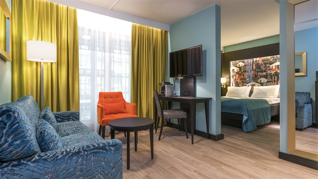 Blå sofa, orange lenestol, skrivebord, smart TV, speil og dobelseng på superiorrom på Thon Hotel Linne