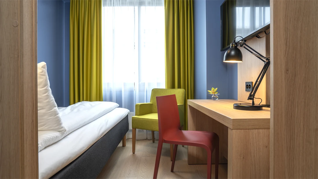 Enkelt seng, blå vegger, gul stol og gule gardiner, skrivebord og TV på enkeltrom på Thon Hotel Astoria