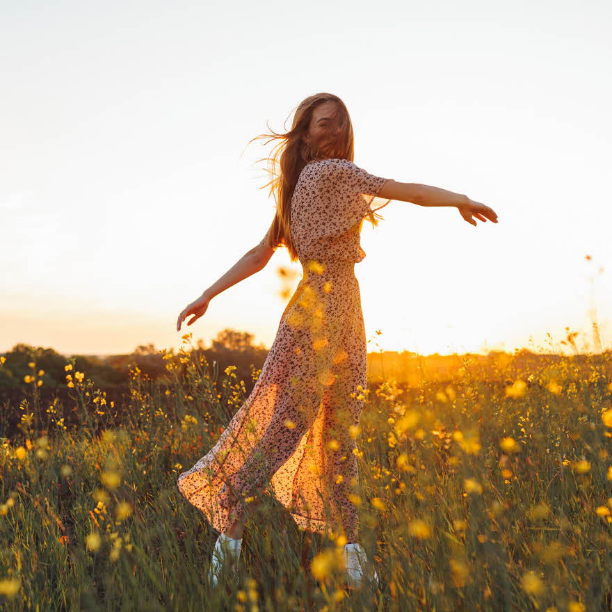Dame med lang blomstrete kjole danser i gul blomstereng i vårsol