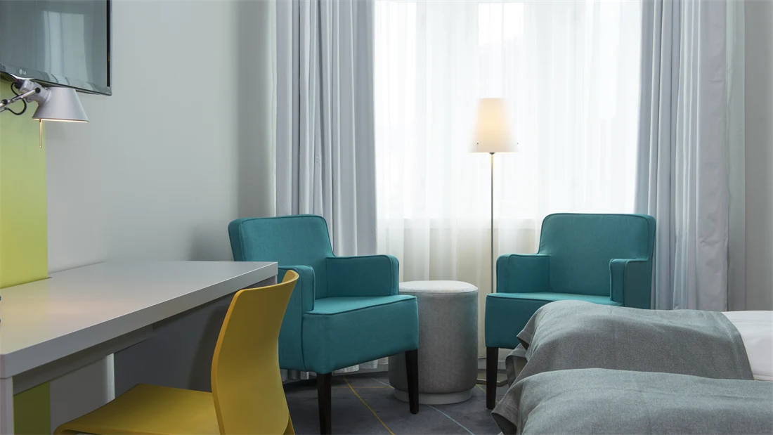 Nærbilde av Standard Twin Room på Thon Hotel Trondheim. Grått skrivebord, gul kontorstol, to turkise lenestoler, to enkeltsenger, vegghengt TV. Store vinduer gir mye godt lys.