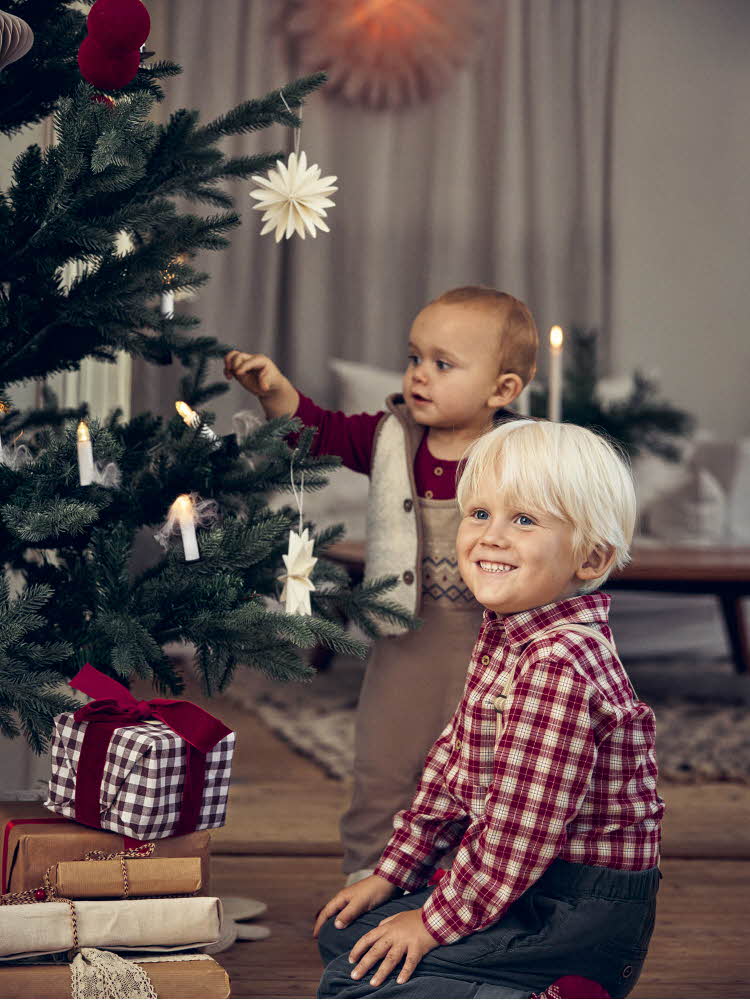 Liten gutt sitter ved juletreet og smiler, baby står i bakgrunnen og tar på en gren.