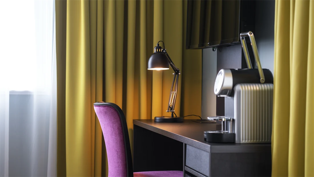 Tv, skrivebord, rosa stol, kaffemaskin, gule gardiner, vindu