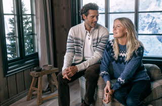 En mann og en dame i ullklær sitter inne med utsikt mot snødekte fjell.