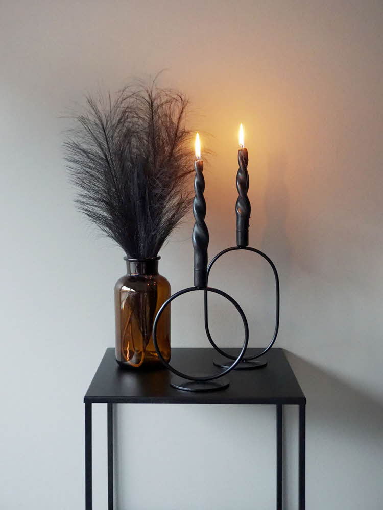 Vase med strå og svarte lysestaker med brennende lys på et svart bord.